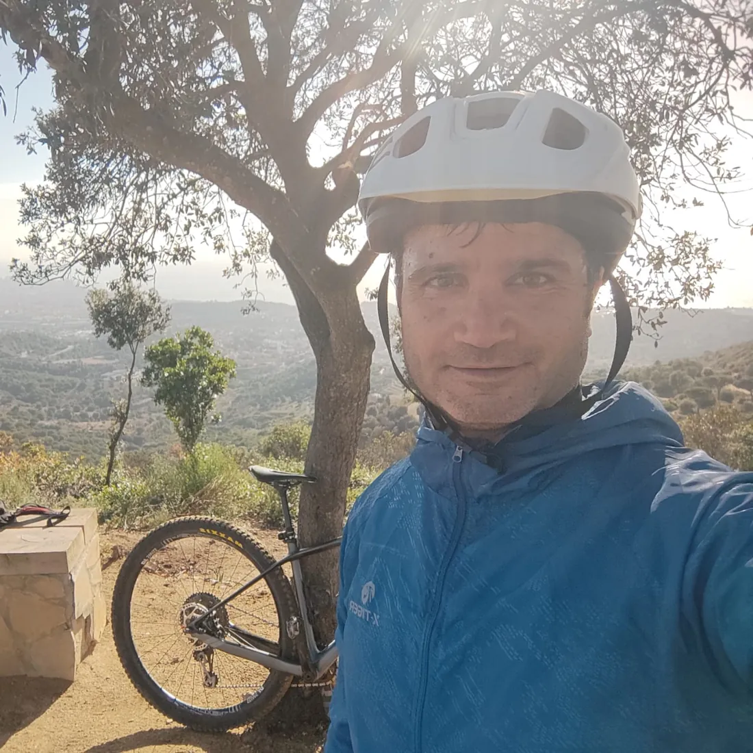 Jordi, autor de ZonaMTB, con casco y chaqueta azul junto a su bici de montaña apoyada en un árbol, con un valle al fondo